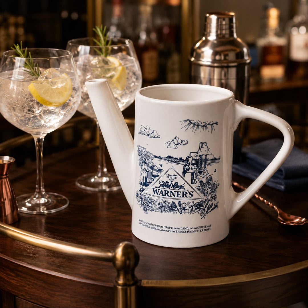 A white ceramic novelty pitcher styled on a dark wooden bar cart, featuring a blue vintage-style illustration of Warner's saloon and a cowboy. Two cocktail glasses with lemon and rosemary are in the background on a blurred bar counter.