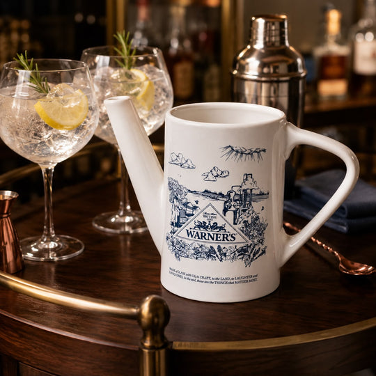 A white ceramic novelty pitcher styled on a dark wooden bar cart, featuring a blue vintage-style illustration of Warner's saloon and a cowboy. Two cocktail glasses with lemon and rosemary are in the background on a blurred bar counter.