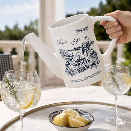 A white ceramic pitcher with a blue 'The Warners' movie illustration, being poured with water from a hand held by a person, set on an outdoor patio next to two glasses of iced tea with lemon.