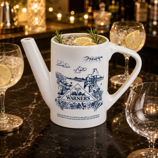 A white ceramic mug styled as a teapot with a blue illustrated print of the Warners movie logo, filled with a cocktail and garnished with rosemary and lemon slices, set on a dark marble bar counter with wine glasses and warm bokeh lights in the background.