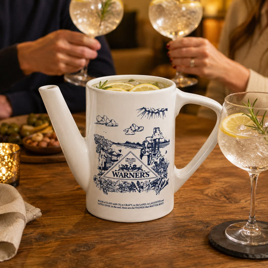 A white ceramic teapot with blue illustrated graphics and the brand name 'WARNERS' on the front, filled with a sparkling drink and lemon slices, set on a wooden table in a warm, social setting with people holding glasses in the background.