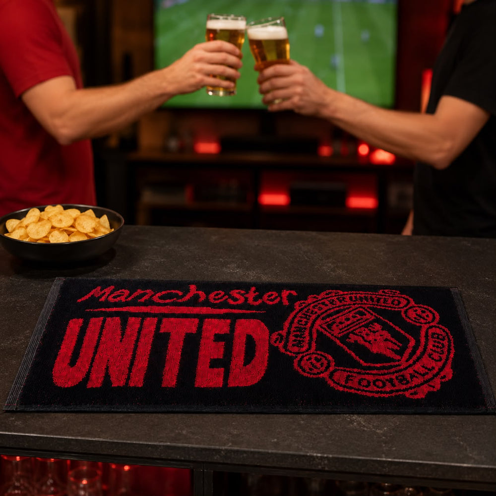 A Manchester United themed bar mat on a dark bar counter, with two people in the background clinking beer glasses and a bowl of potato chips on the counter.