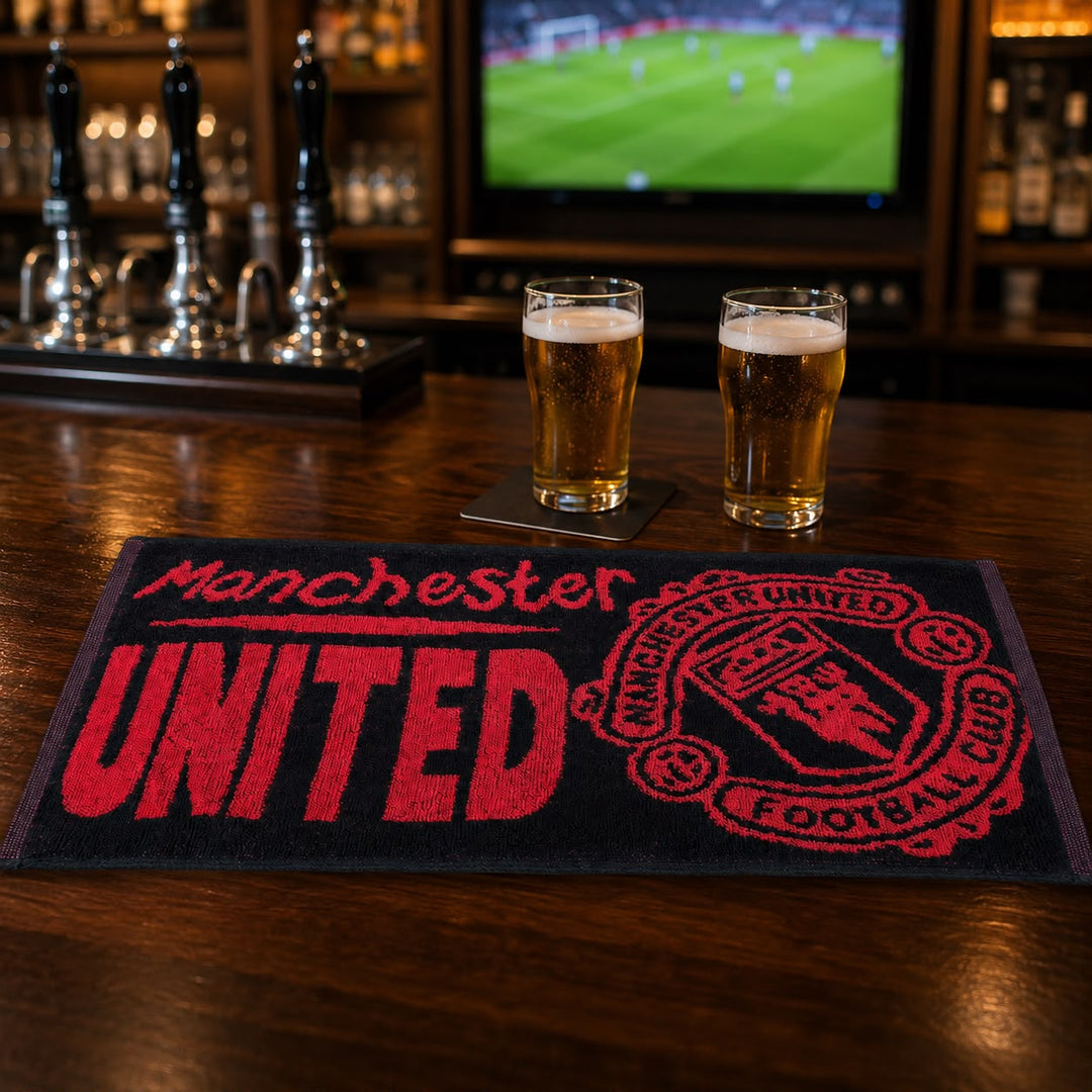 A Manchester United themed bar mat on a wooden bar table, with two glasses of beer on coasters and a blurred TV screen in the background.