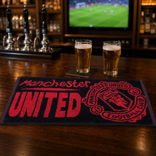 A Manchester United themed bar mat on a wooden bar table, with two glasses of beer on coasters and a blurred TV screen in the background.