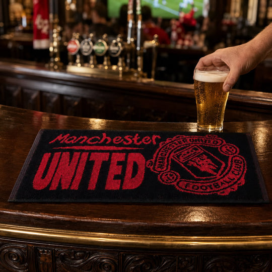 A black Manchester United bar mat with red embroidered-style lettering and crest sits on a polished wooden bar counter. A hand holding a pint of beer is visible at the right edge, and the bar interior is softly blurred in the background.