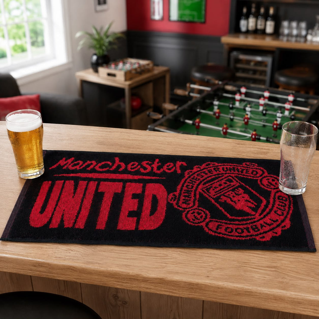 A black Manchester United football club mat with red lettering and crest, displayed on a wooden bar table in a bar setting with foosball and stools in the background.