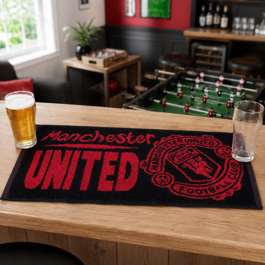A black Manchester United football club mat with red lettering and crest, displayed on a wooden bar table in a bar setting with foosball and stools in the background.