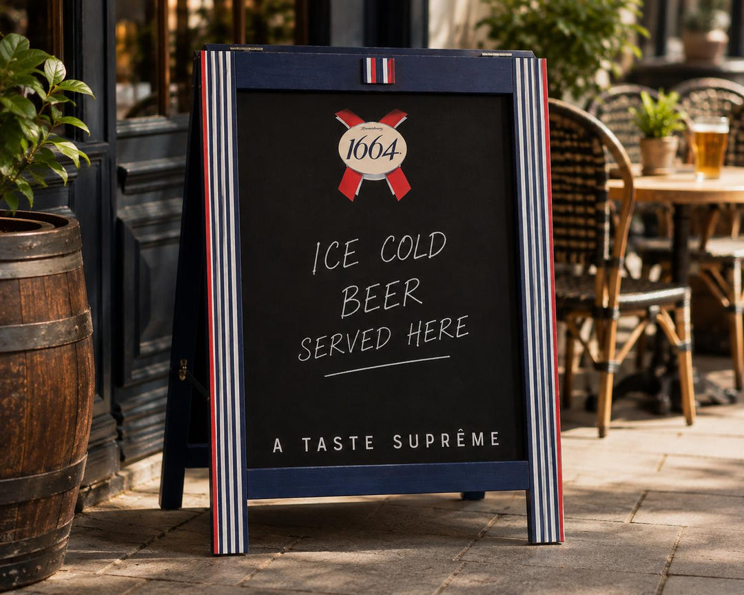 A black A-frame sidewalk sign with red and white striped borders stands on a stone patio outside a restaurant. The sign reads '1664' with a ribbon motif, 'ICE COLD BEER SERVED HERE,' and 'A TASTE SUPRÈME.